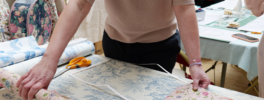 Une personne mesure et dispose des fleurs et du tissu « Toile de Jouy » sur une table, avec des ciseaux à portée de main et d’autres rouleaux de tissu ainsi que du matériel de loisirs créatifs à l’arrière-plan, créant ainsi une charmante ambiance d’atelier pour adultes, éclairée par une petite lampe. - Musée de la Toile de Jouy