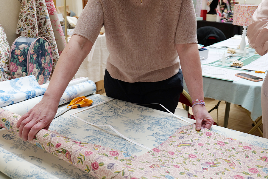 Une personne mesure et dispose des fleurs et du tissu « Toile de Jouy » sur une table, avec des ciseaux à portée de main et d’autres rouleaux de tissu ainsi que du matériel de loisirs créatifs à l’arrière-plan, créant ainsi une charmante ambiance d’atelier pour adultes, éclairée par une petite lampe. - Musée de la Toile de Jouy
