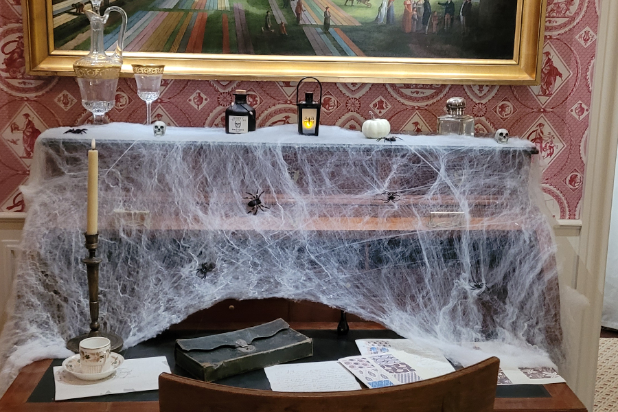 Un bureau recouvert de fausses toiles d'araignées, agrémenté d'araignées, de bougies, de crânes, d'une lanterne, d'une citrouille blanche et de lettres manuscrites, crée une ambiance effrayante d'Halloween qui rappelle un atelier créatif — une source d'inspiration idéale pour les fêtes de la Toussaint. - Musée de la Toile de Jouy
