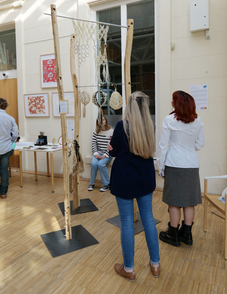 Deux personnes admirent des œuvres d’art en macramé suspendues à des cadres en bois, tandis qu’une autre personne est assise à proximité. L’exposition, organisée dans le cadre des Journées européennes du patrimoine, se tient à l’intérieur : on y voit des œuvres accrochées aux murs et une table à l’arrière-plan. Elle met à l’honneur le patrimoine à travers des événements culturels uniques. - Musée de la Toile de Jouy