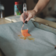 Dans le cadre d'une initiation à l'atelier enfants, une personne utilise un pinceau en silicone pour étaler de l'huile sur du papier sulfurisé recouvrant une plaque de cuisson. Seules ses mains et une partie de son torse sont visibles. - Musée de la Toile de Jouy