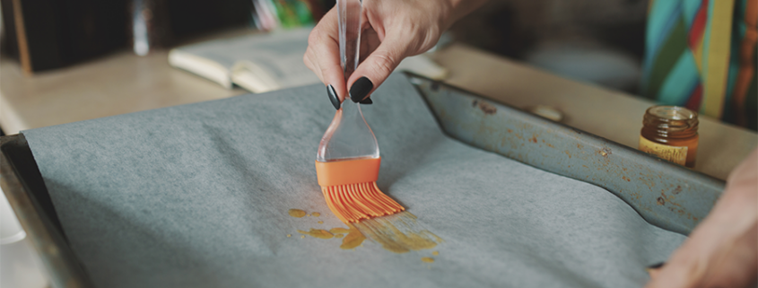 Dans le cadre d'une initiation à l'atelier enfants, une personne utilise un pinceau en silicone pour étaler de l'huile sur du papier sulfurisé recouvrant une plaque de cuisson. Seules ses mains et une partie de son torse sont visibles. - Musée de la Toile de Jouy
