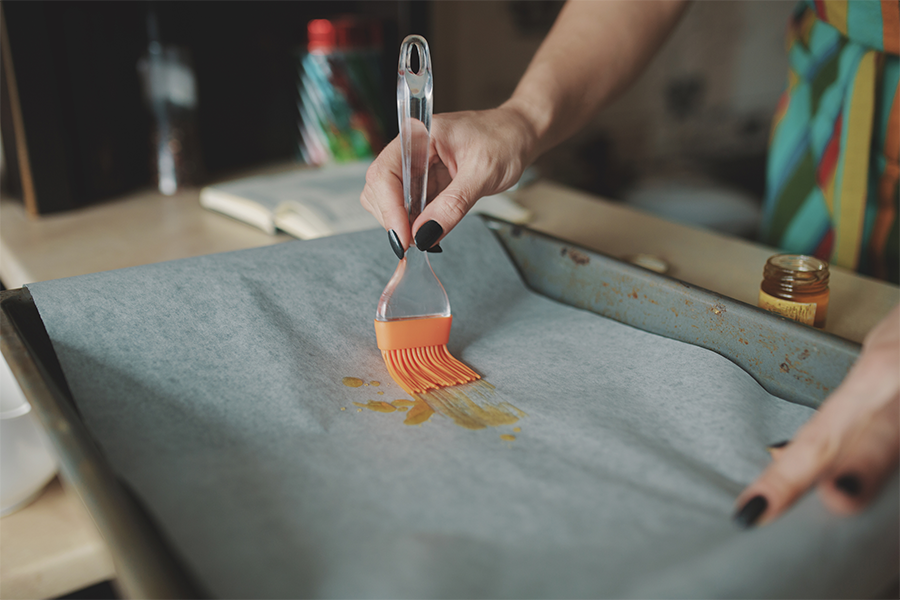 Dans le cadre d'une initiation à l'atelier enfants, une personne utilise un pinceau en silicone pour étaler de l'huile sur du papier sulfurisé recouvrant une plaque de cuisson. Seules ses mains et une partie de son torse sont visibles. - Musée de la Toile de Jouy