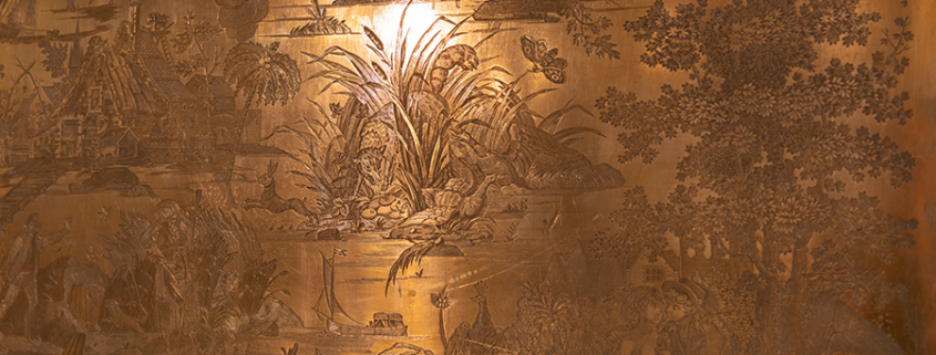 Panneau décoratif aux reflets dorés, orné de scènes naturelles gravées représentant notamment des plantes, des arbres, un chalet, une rivière, des bateaux et des personnages, avec des détails minutieux et un éclat doux se reflétant sur la surface — un ajout remarquable à toute collection permanente ou un élément d'élégance qui mettra en valeur votre visite guidée. - Musée de la Toile de Jouy