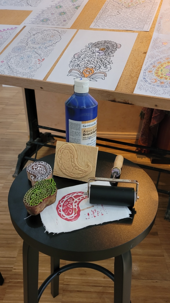 Matériel d'impression au bloc posé sur un tabouret, comprenant de l'encre, un rouleau, des tampons et du tissu imprimé, tandis qu'à l'arrière-plan, les impressions terminées sèchent sur une table. - Musée de la Toile de Jouy