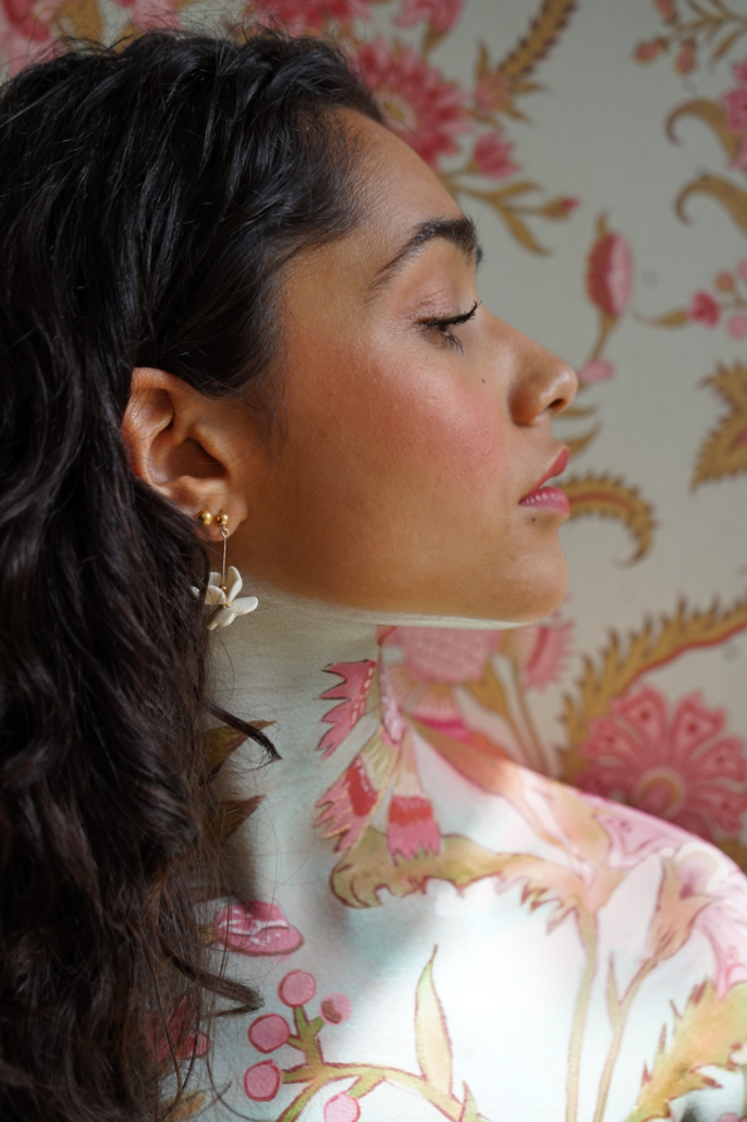 Une femme aux cheveux bruns ondulés, portant des boucles d'oreilles à motifs floraux, pose de profil devant un fond de papier peint aux motifs floraux très travaillés ; son cou et ses épaules sont ornés de motifs floraux assortis — un hommage artistique à l'esprit du patrimoine célébré lors des Journées européennes du patrimoine. - Musée de la Toile de Jouy
