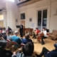 Un petit ensemble joue de la flûte et du violoncelle dans un salon de musique devant un public assis dans une grande salle. - Musée de la Toile de Jouy