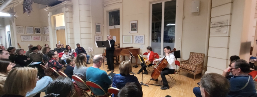Un petit ensemble joue de la flûte et du violoncelle dans un salon de musique devant un public assis dans une grande salle. - Musée de la Toile de Jouy